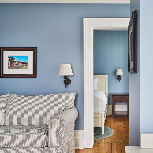 A living room with a striped sofa, wall art, and a lamp, adjacent to a bedroom featuring a bed, nightstand, and lamp.