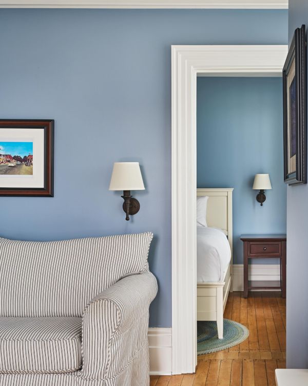 A living room with a striped sofa, wall art, and a lamp, adjacent to a bedroom featuring a bed, nightstand, and lamp.