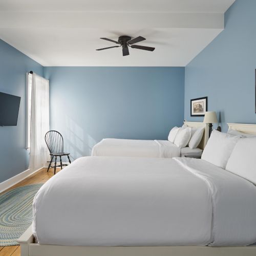 A cozy room with two beds, blue walls, a ceiling fan, a chair, and a wall-mounted TV. There's also a framed picture and a round rug.