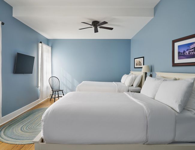 A cozy room with two beds, blue walls, a ceiling fan, a chair, and a wall-mounted TV. There's also a framed picture and a round rug.