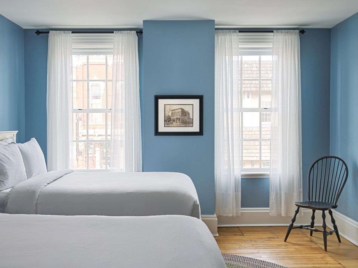 A bedroom with two beds, a wooden chair, picture frame, and two windows with sheer curtains. The walls are painted light blue.