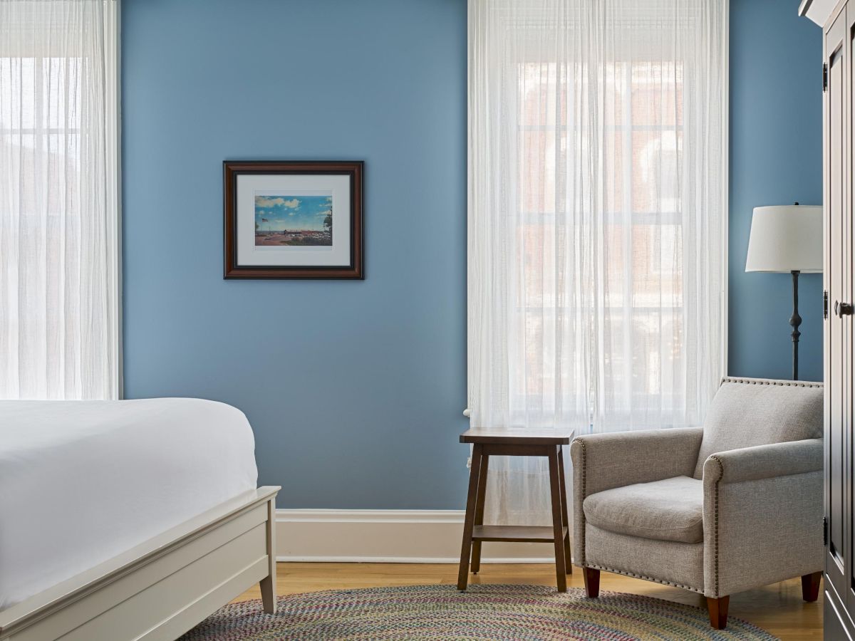 A cozy bedroom with blue walls, a bed, armchair, small table, and lamp by two windows with sheer curtains, and a framed picture.
