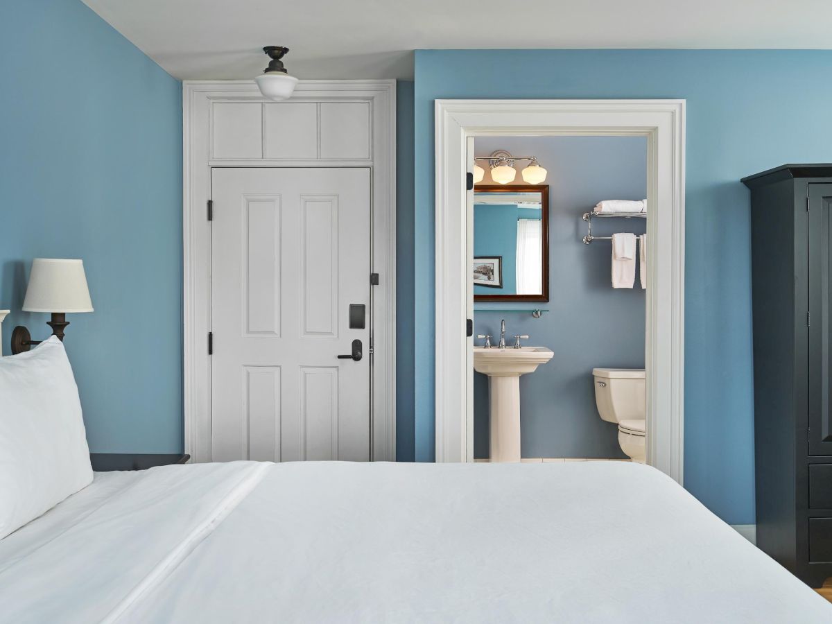 A bedroom with blue walls, white bed, black wardrobe, and a view into a bathroom with a sink and toilet.