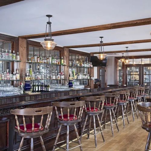 The image shows a rustic bar with wooden stools, a well-stocked shelf with bottles, and hanging lights, creating a cozy atmosphere.