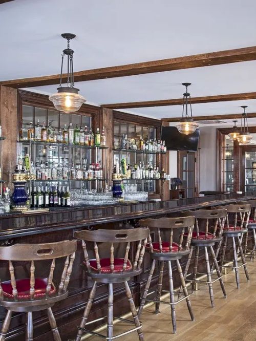 The image shows a rustic bar with wooden stools, a well-stocked shelf with bottles, and hanging lights, creating a cozy atmosphere.