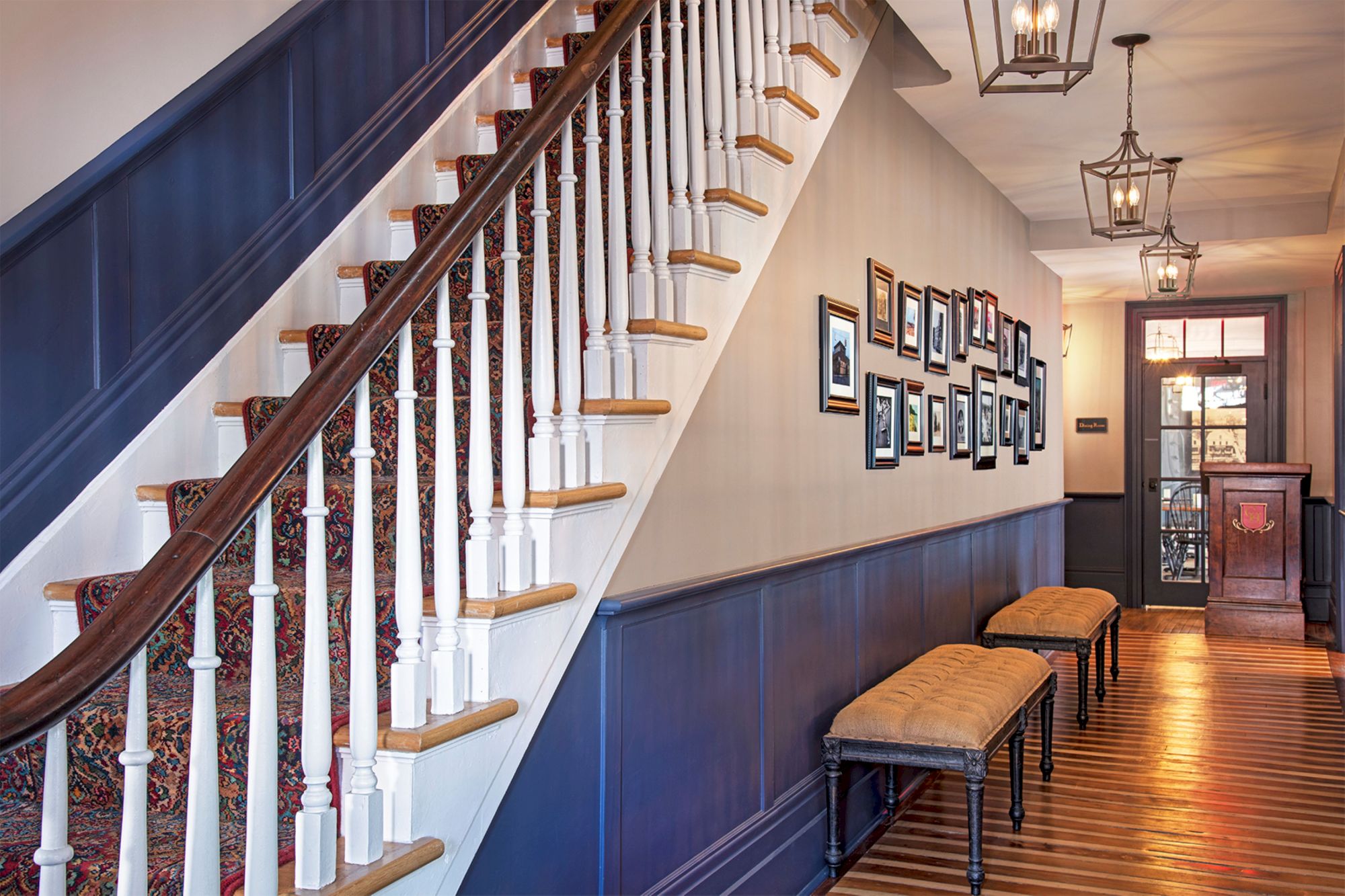 A staircase with carpet, picture frames on the wall, benches, and overhead lighting in a hallway with wooden flooring and painted paneling.
